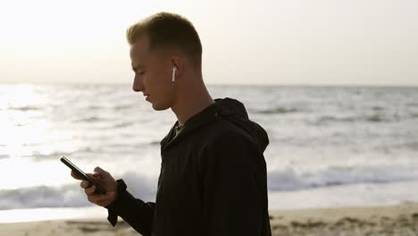 portrait of a young man who listens to music on headphones using his phone and walks along the beach in the morning