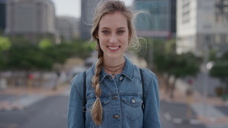 ritratto bella giovane donna bionda studentessa sorridente in città godendo di uno stile di vita urbano indipendente vento che soffia i capelli indossando una giacca denim slow motion