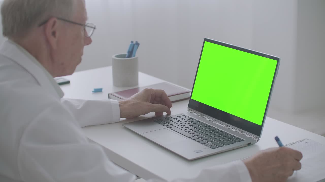 un anciano médico está leyendo un artículo en internet, mirando la pantalla de la tecla de croma de la computadora portátil y escribiendo