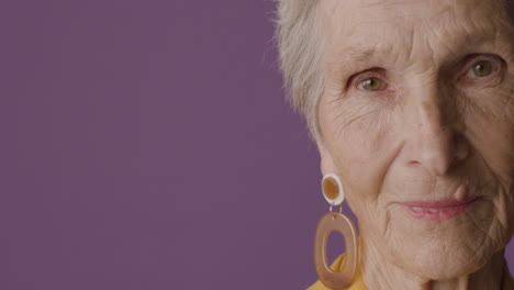 Close-Up-View-Of-Senior-Woman-Face-With-Short-Hair-And-Green-Eyes-Wearing-Earrings-Posing-And-Smiling-At-Camera-On-Purple-Background