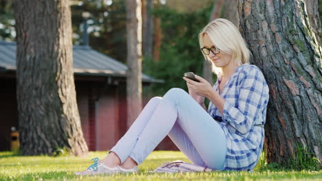 a young woman is using a smartphone sits on the grass under a tree in the backyard of the house 4k v