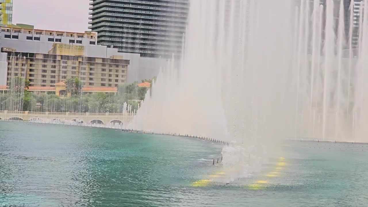 Famoso espectáculo de fuentes de Las Vegas con chorros de agua en arco, reflejos brillantes en el lago, horizonte de hoteles, detalles de puente de piedra, árboles y ambiente de resort turístico diurno