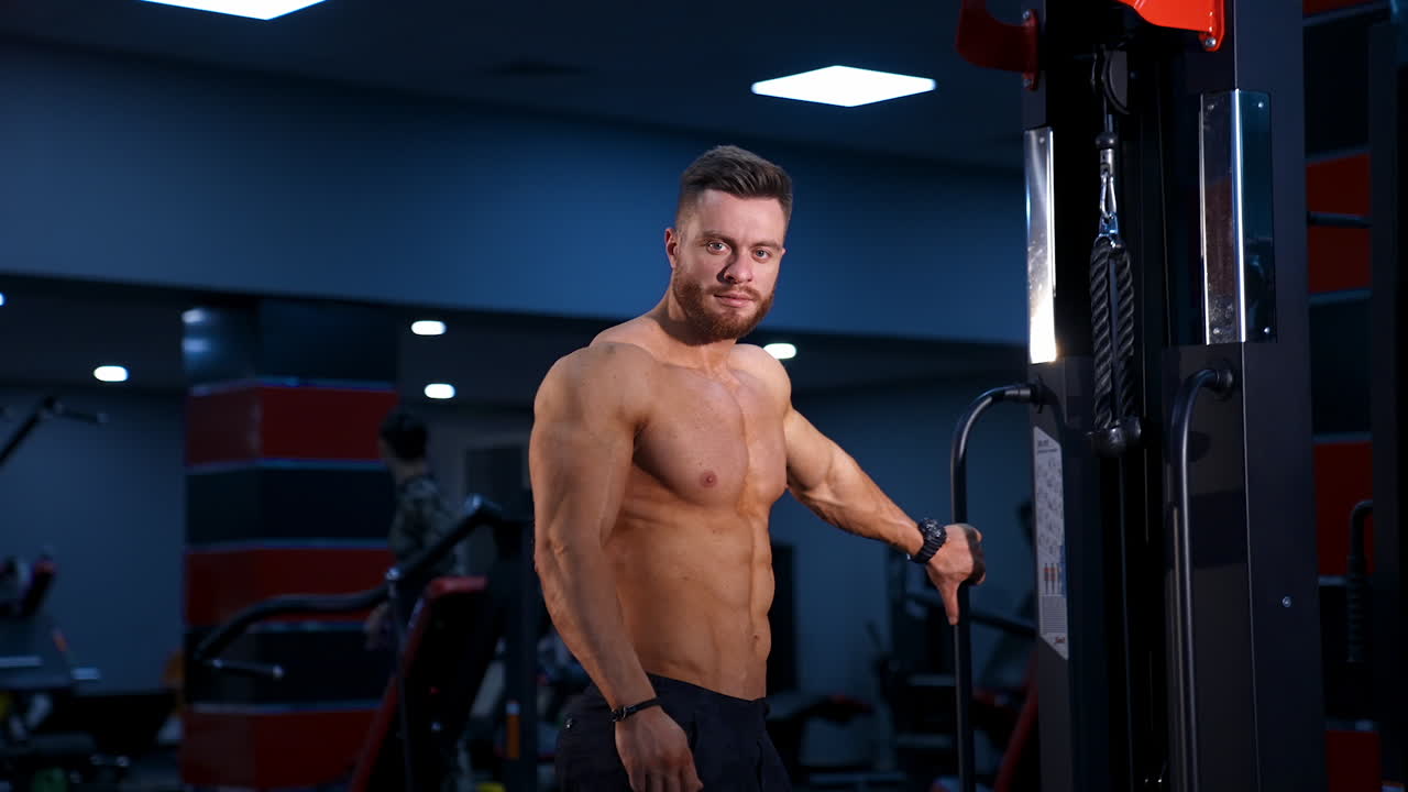 Bodybuilder is posing. Beautiful sporty guy male power. Fitness muscled man without shirt on dark gym background. Slow motion.