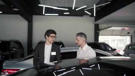 a confident brunette guy with glasses and a business suit an assistant at a car showroom tells a middle-aged man in a white shirt about modern cars at the car showroom