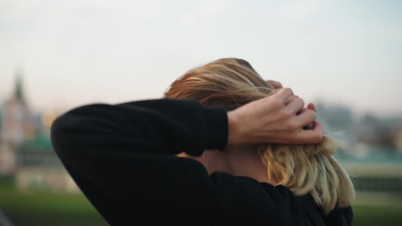 Giovane uomo bianco di profilo con skyline cittadino, mani che passano tra i capelli biondi, da un punto di osservazione su un tetto, felpa nera casual, postura riflessiva che suggerisce un pendolare o un artista, lontane guglie di chiese e sfondo sfocato.