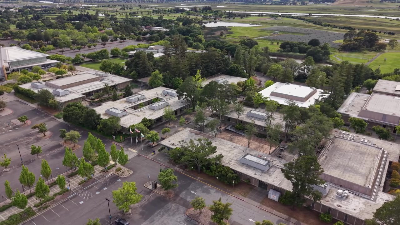 Expansive aerial view of the Napa Valley College campus, showcasing its modern architecture, empty parking lots, and surrounding lush green landscape on a cloudy day in California