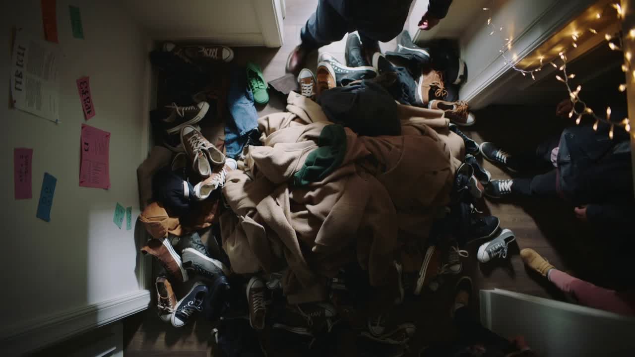 Piedi che entrano dalla porta a destra fanno muovere le persone nel corridoio, aggirando un mucchio di cappotti, scarpe e luci.