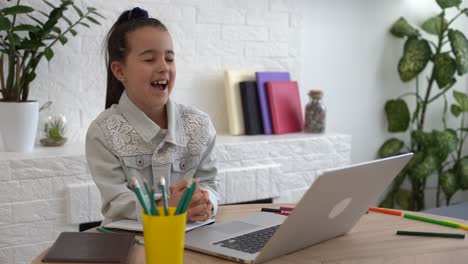 Closeup-photo-of-little-pretty-pupil-school-talk-skype-notebook-online-lesson-video-call-sit-desk,-distance,-classmates-quarantine-study-living-room-indoors