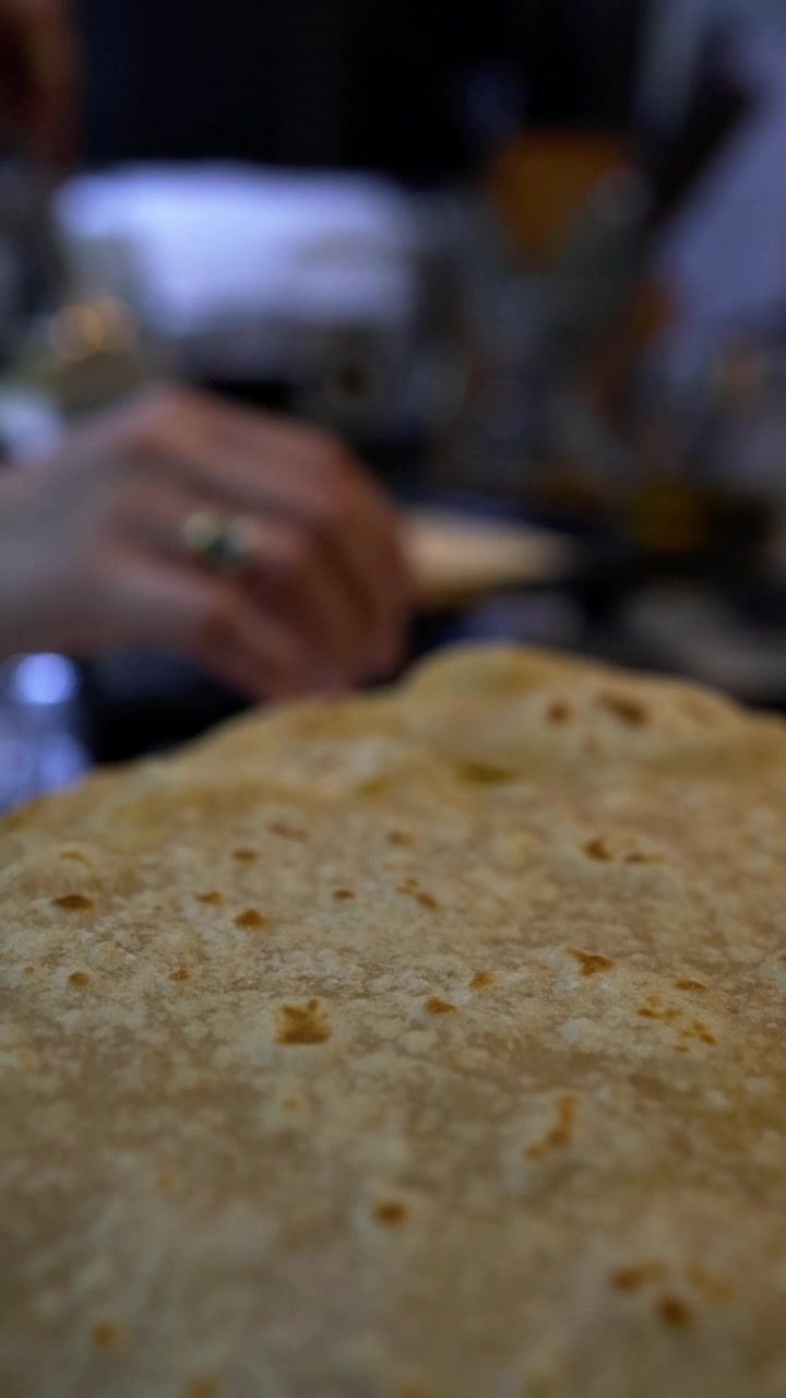 Close-up of freshly cooked chapati flatbread with visible brown spots on surface, hands and cooking utensils blurred in background. Traditional Indian food preparation in home kitchen setting.