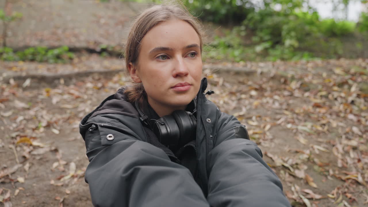 Young white woman resting on forest floor among fallen leaves, wearing dark jacket and headphones, eyes closing then opening to steady gaze, hands stretching then relaxing, mood calm and reflective