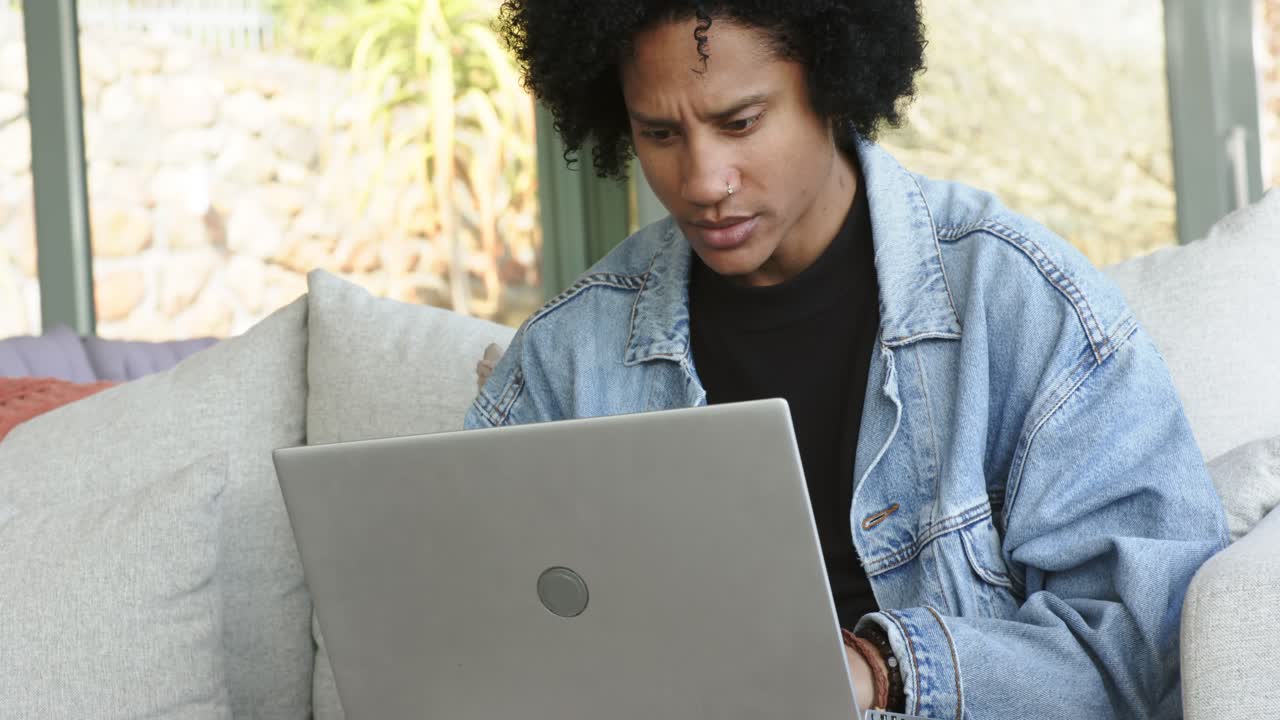 Mid adult African American man placing laptop on lap, remotely typing on sofa by glass door