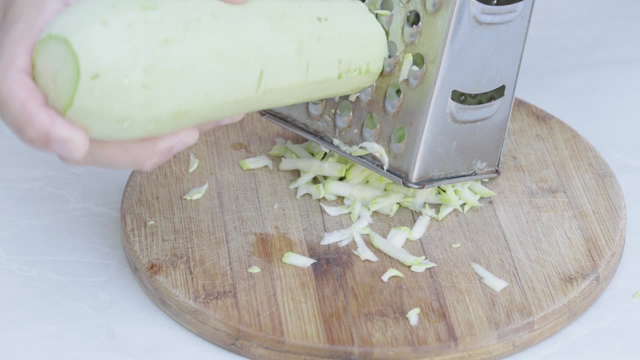 Zucchine grattugiate su grattugia metallica sopra un tagliere di legno, in primo piano, durante la preparazione del cibo.