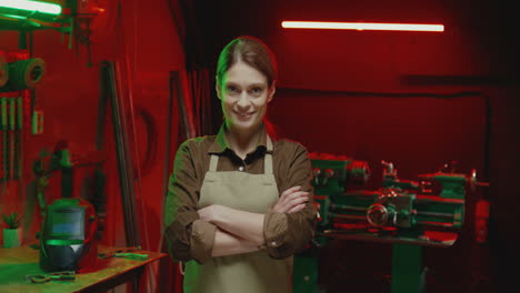 portrait of young woman in apron looking at camera with arms crossed in metal workshop