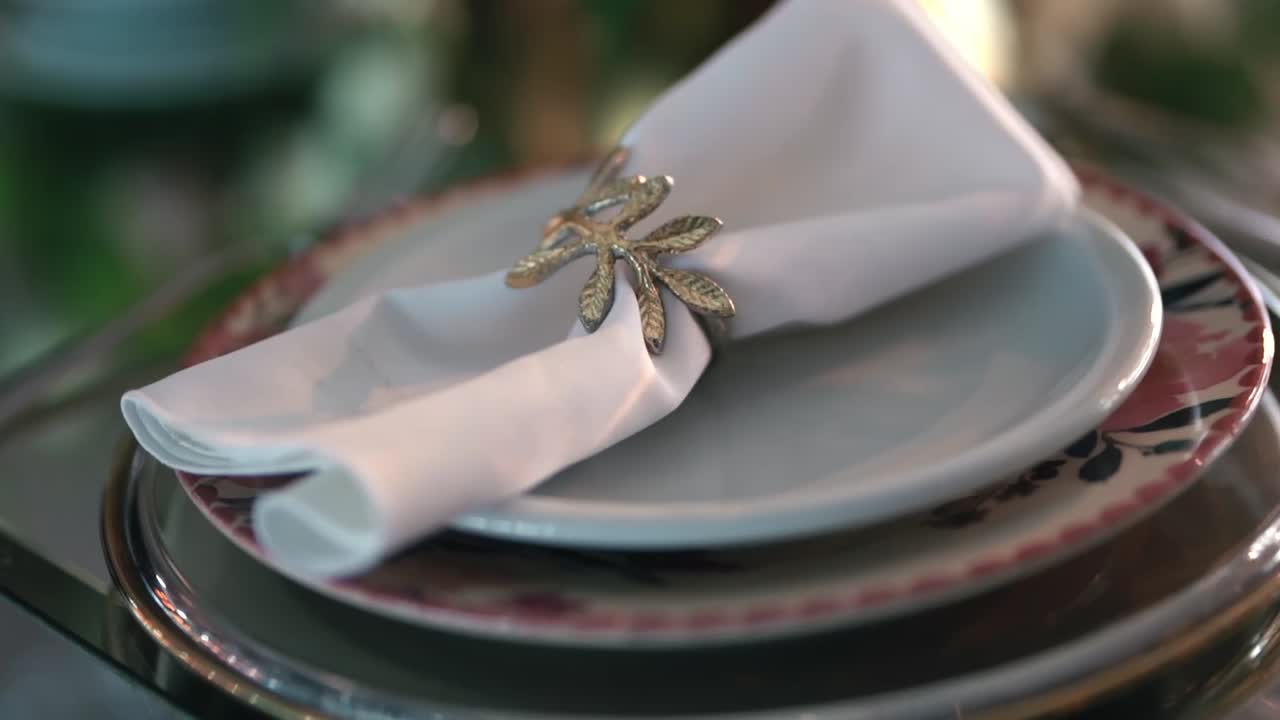 Ripresa fluida e lenta con carrello in avvicinamento che mette a fuoco un tovagliolo bianco con un anello decorato a forma di foglia su un piatto di ceramica, mostrando l'elegante decorazione della tavola di un ricevimento nuziale di lusso
