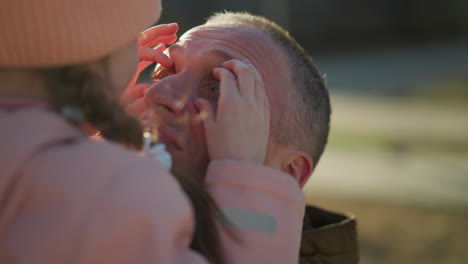 a joyful little girl in a pink cap and jacket is being carried by a man as she playfully touches his eyes with her hands. the intimate moment is filled with warmth and happiness