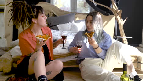 two happy girls drinking wine and sharing pizza while sitting on the floor at home 1