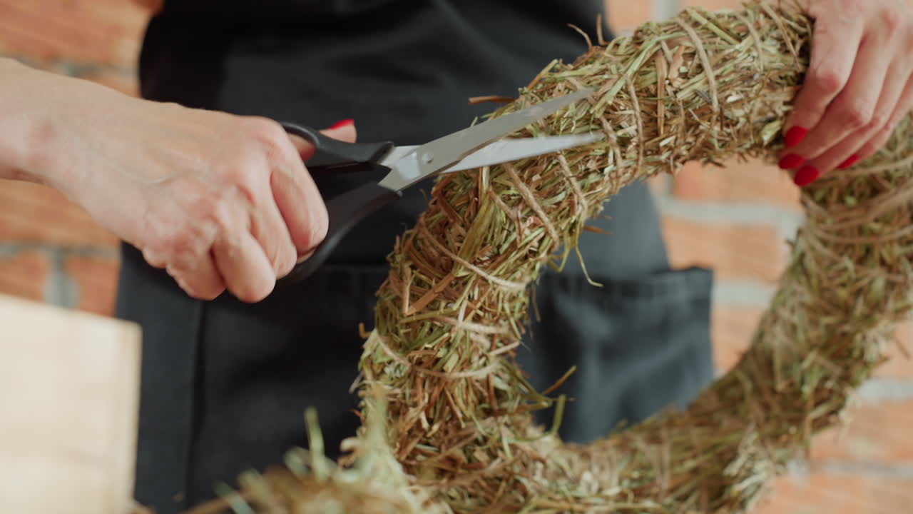 Closeup of woman using scissors to trim straw wreath on wooden table, focusing on precise crafting process, handmade decoration and attention to artisan details in floral design preparation