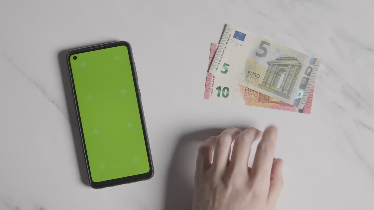 fotografía aérea de una persona que toca los dedos junto a billetes de 10 y 5 euros y un teléfono móvil con pantalla verde