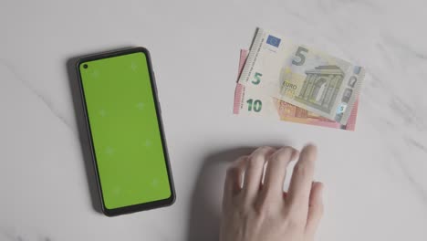 Overhead-Currency-Shot-Of-Person-Tapping-Fingers-Next-To-10-And-5-Euro-Notes-And-Green-Screen-Mobile-Phone