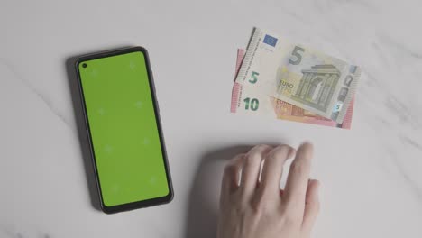 fotografía aérea de una persona que toca los dedos junto a billetes de 10 y 5 euros y un teléfono móvil con pantalla verde