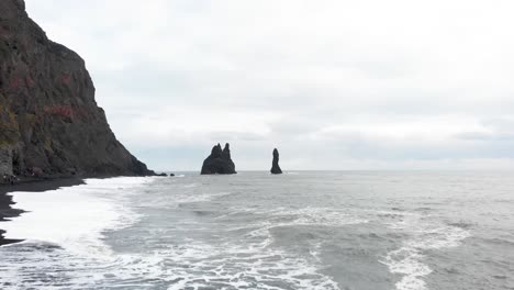 Olas-Espumosas-Golpeando-La-Playa-De-Arena-Negra-De-Reynisfjara,-Columnas-De-Roca-Más-Allá