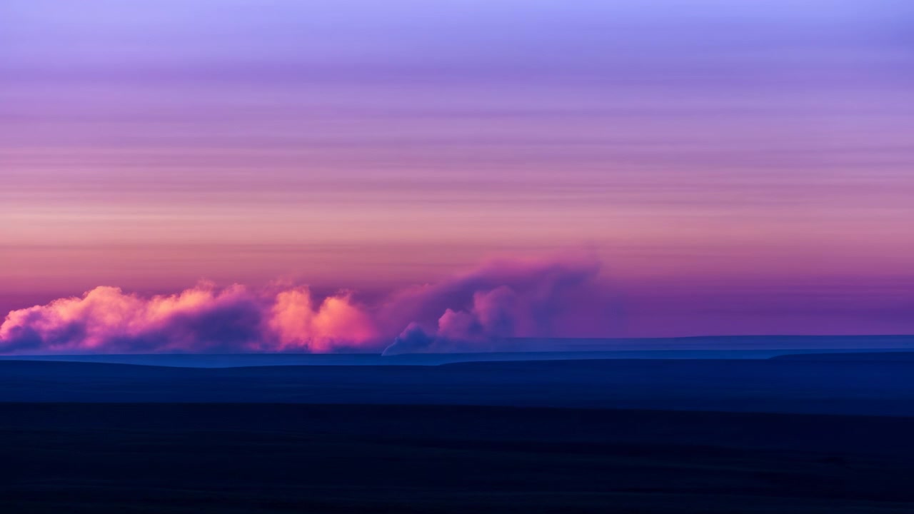 Nebbia bassa o nuvola si estende lungo l'orizzonte sopra pianure in silhouette, mentre una calda luce rosa e arancione a sinistra sfuma in toni più freddi di viola e blu, rivelando un'atmosfera calma e stratificata.
