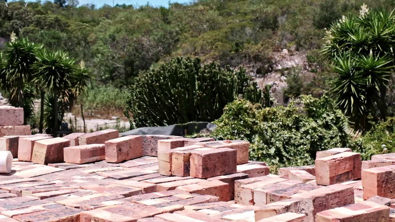 A pile of clay bricks stacked up on a building site with a beautiful, lush green view behind