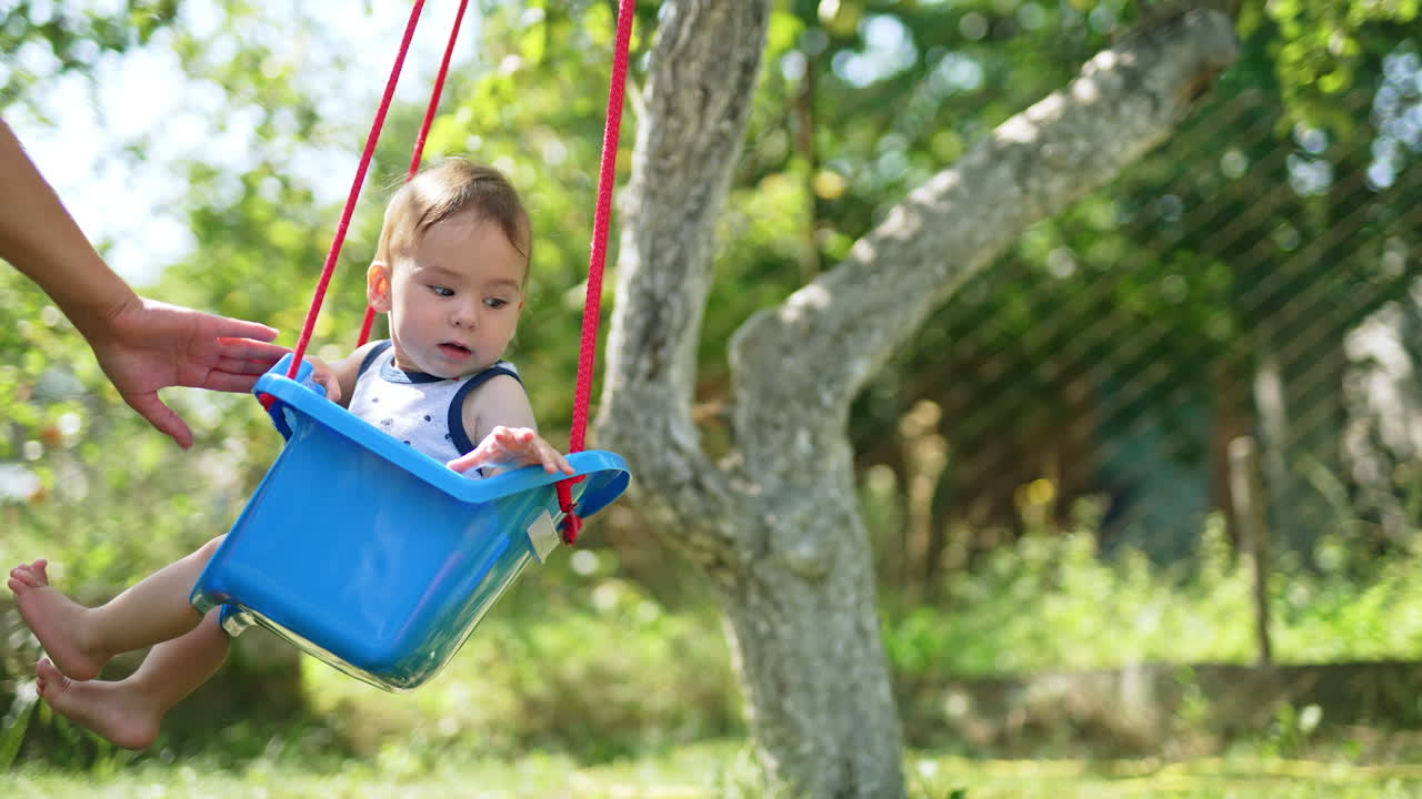 Parent's hand pushing the blue swing with cute baby sitting inside. Fun time with kid in the garden in summer. Nature backdrop.