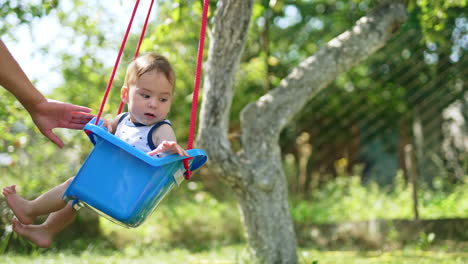 parent's hand pushing the blue swing with cute baby sitting inside. fun time with kid in the garden in summer. nature backdrop.
