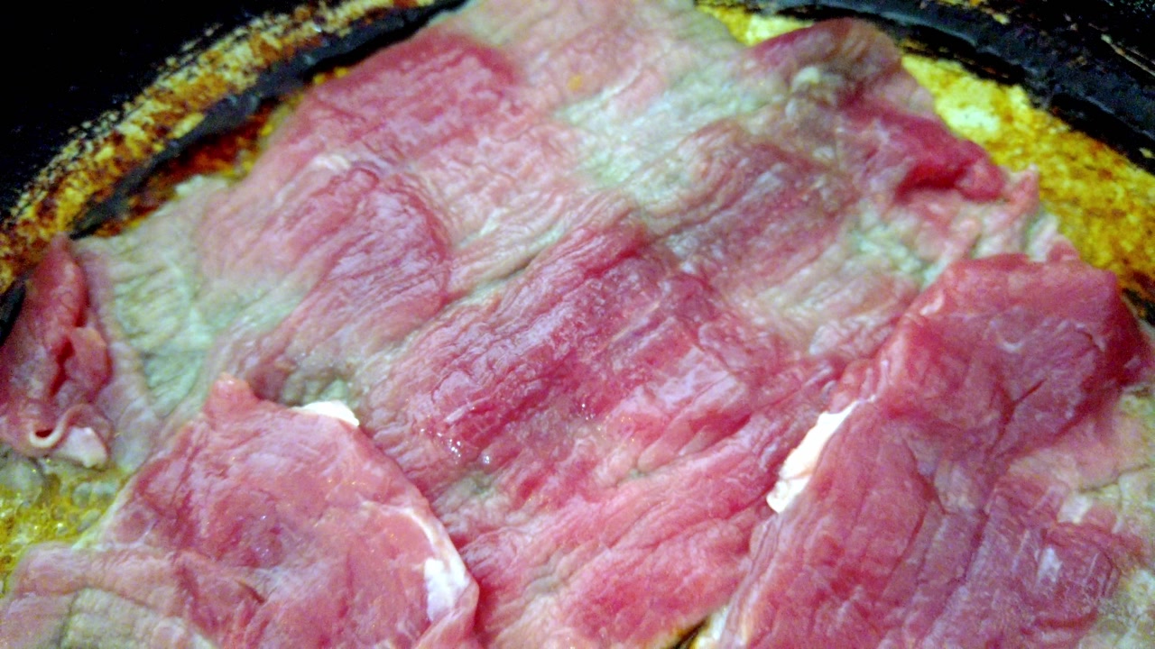 Close-up of raw beef steak being cooked on a hot pan with sizzling oil.