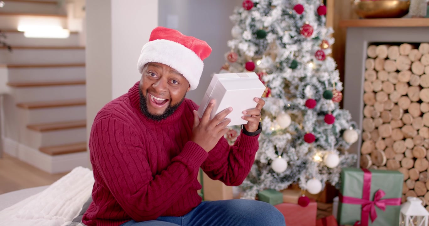 Afroamerikansk mann sitter på sofa nær juletre, holder hvit eske, undersøker og viser glede