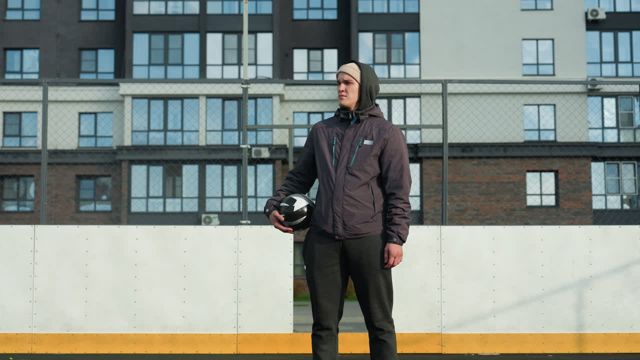 Youngster in hoodie and beanie holding ball in right hand stands on football pitch with goal post, tall office building in background, and sunlight casting shadow on him