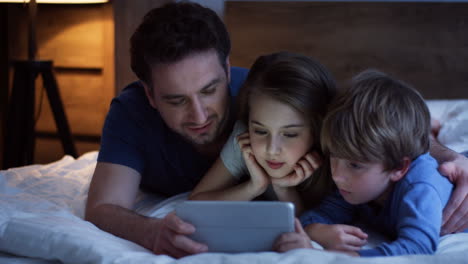 padre caucásico y su pequeño hijo e hija acostados en la cama y viendo algo en la tableta
