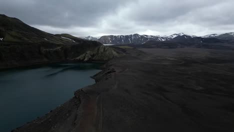 Hnausapollur-aerial-view-in-the-highlands-of-Iceland,-with-a-mountain-covered-in-snow-in-the-background,-different-shades-of-grey-and-green,-and-a-blue-crater-laker-in-a-beautiful-landscape