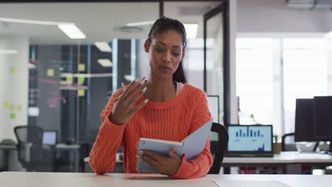 Mixed-race-businesswoman-sitting-at-desk-holding-notebook-having-video-call-gesturing