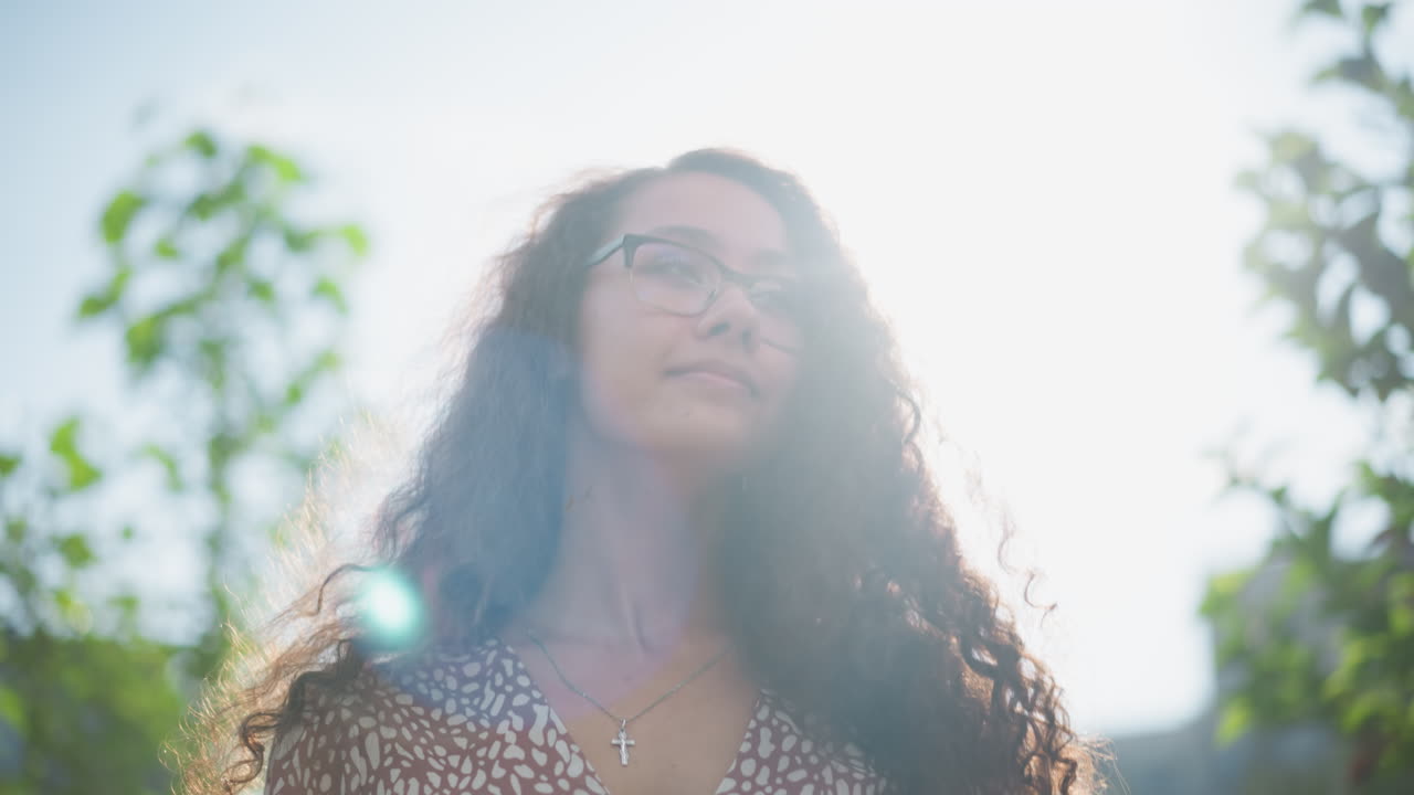 mujer con gafas y colgante de cruz ajusta su cabello al aire libre, sonriendo cálidamente mientras la luz del sol se refleja en su cara, rodeada de vegetación y una suave luz natural