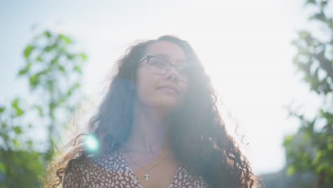 woman with glasses and cross pendant adjusts her hair outdoors, smiling warmly as sunlight reflects off her face, surrounded by greenery and a soft natural light