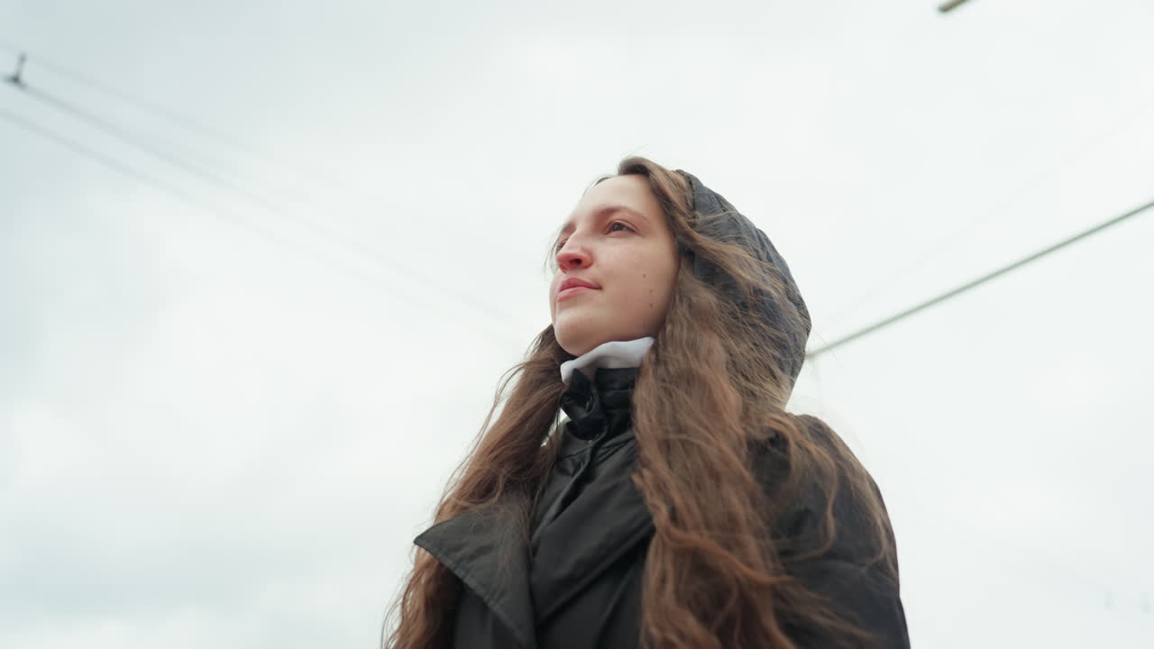 hooded white woman walking urban avenue with long hair blowing, side profile near highrise buildings and tram cables, confident gaze toward skyline, casual coat, morning commute vibe, roles include