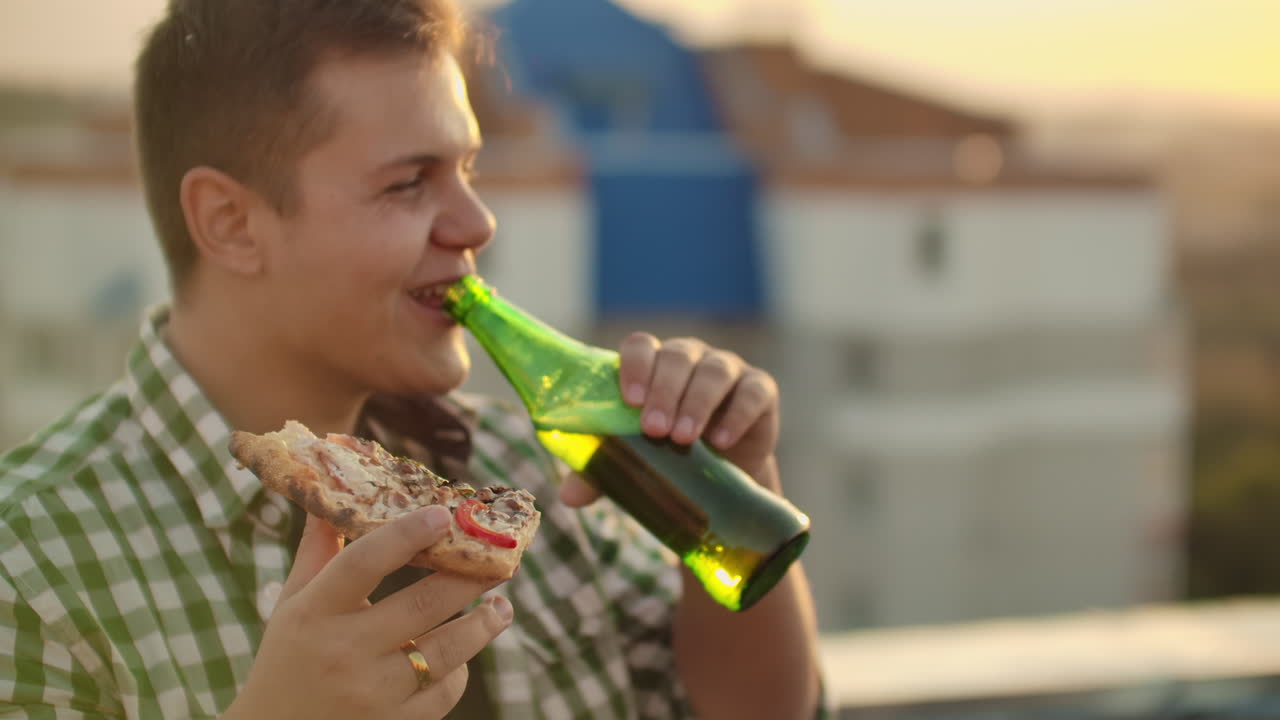 un niño come pedazos de pizza caliente y bebe cerveza en el techo. sonríe y disfruta del momento.