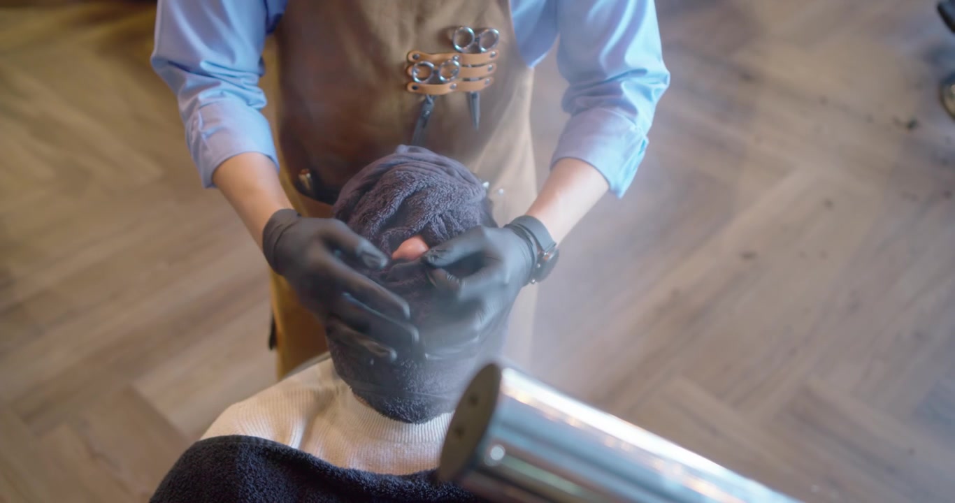 Afro-Amerikaanse mannelijke stylist die een stomende handdoek over een mannelijke klant in de salon legt, ter voorbereiding op het scheren.
