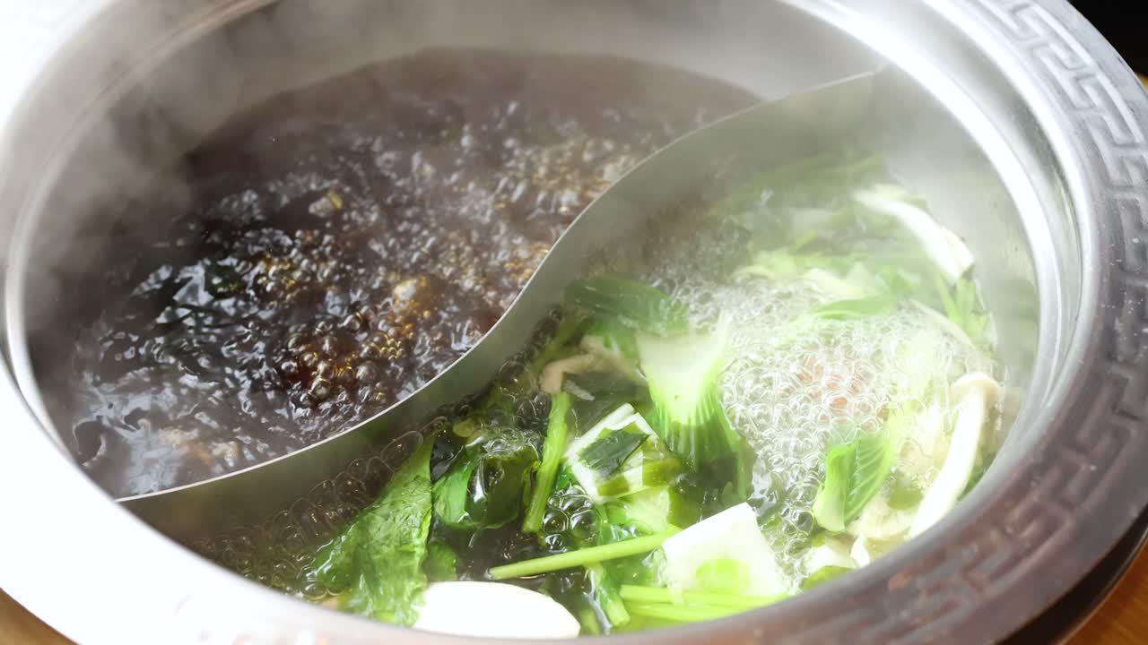 Overhead view of steaming dual hotpot with spicy and clear broth, fresh vegetables, and herbs