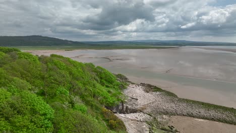 Green-and-rugged-coastline-flyover-towards-cloudy-bright-horizon-on-spring-day-at-Jenny-Brown's-Point,-Silverdale,-Lancashire,-England,-UK