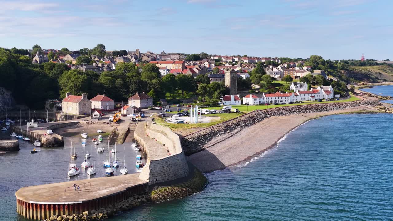 Drone glides above coastal town, revealing harbor, boats, beachfront houses, and scenic shoreline in sunlight
