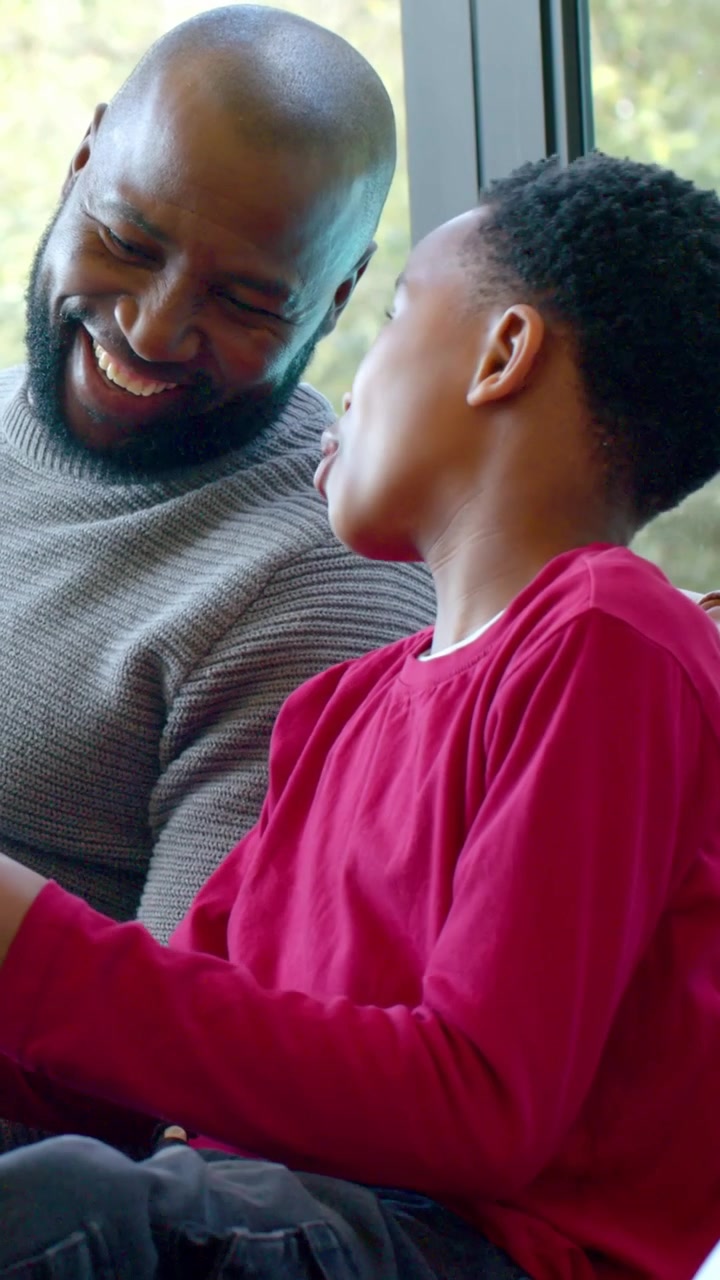 Vertical video: Tapping tablet, boy in red shirt engaging father in gray sweater on couch by window