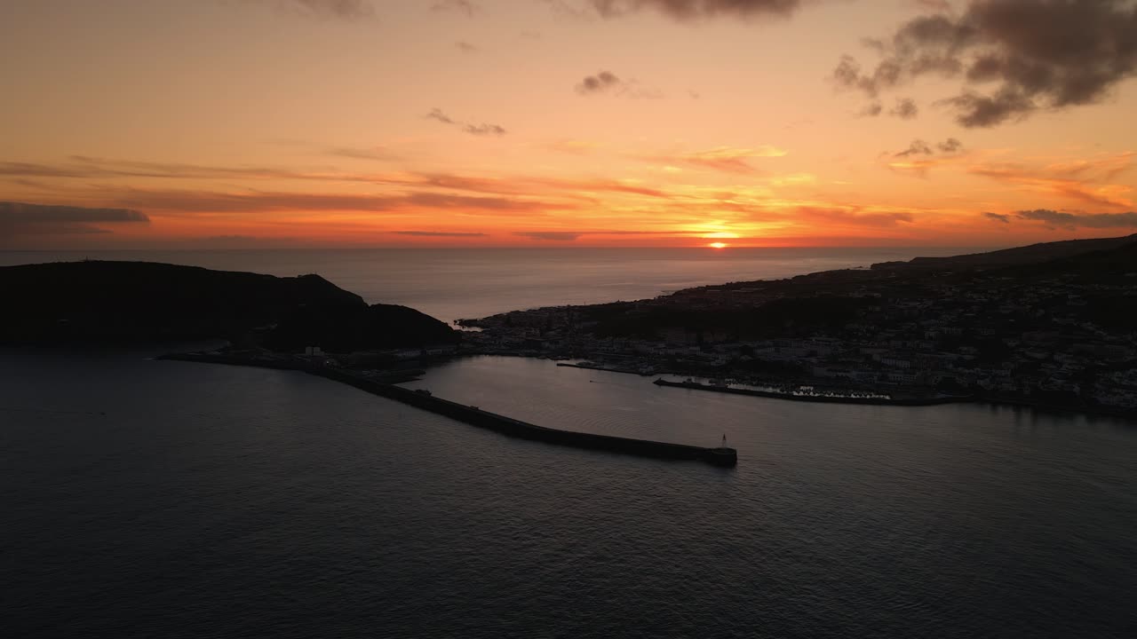 A sweeping view of the city lights beginning to flicker in a coastal town with the last rays of sun in the background