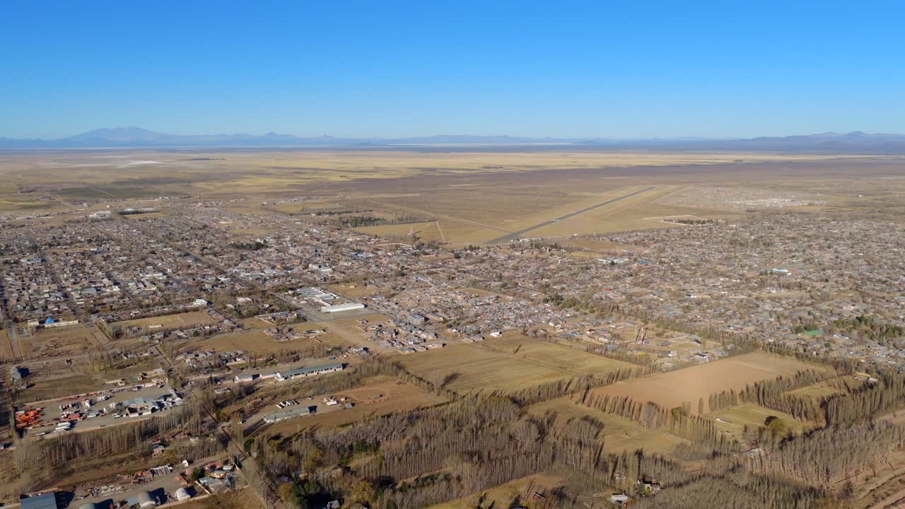 Slow wide drone glide over southern edge of Malargüe in Mendoza province, revealing farmland patterns, town limits and vast arid plains under clear skies with distant mountain silhouettes