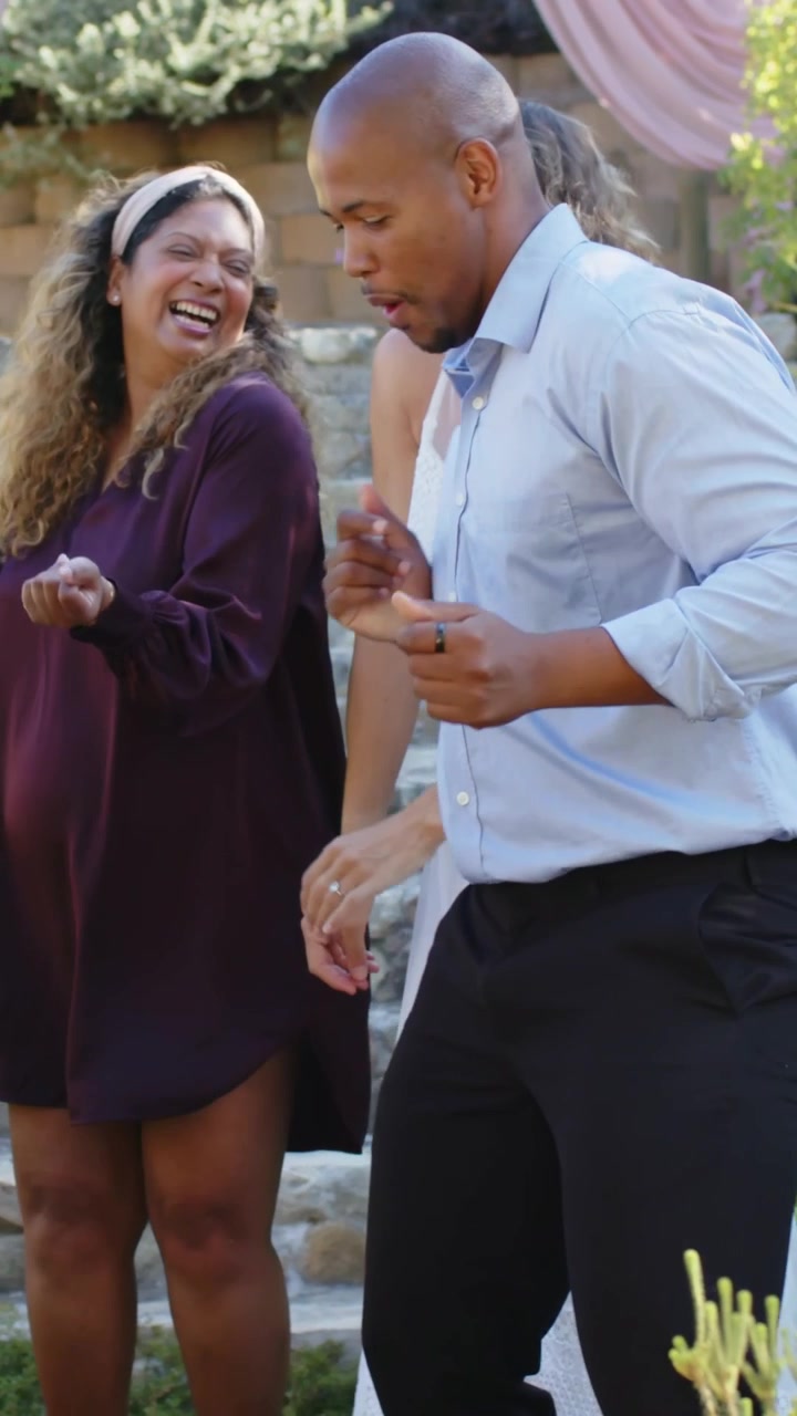 Video verticale: uomo che balla in camicia facendo passi mentre una donna in abito bianco alza le braccia a una festa in giardino.