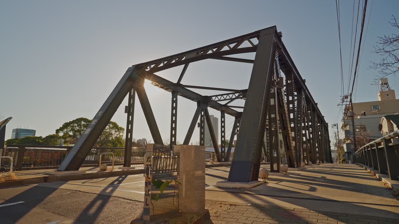 Daiei Bridge's aged dark iron truss rises against a pale hazy sky at golden hour in Tokyo, with trees and city buildings in background, captured as a static low-angle shot from the bridge approach