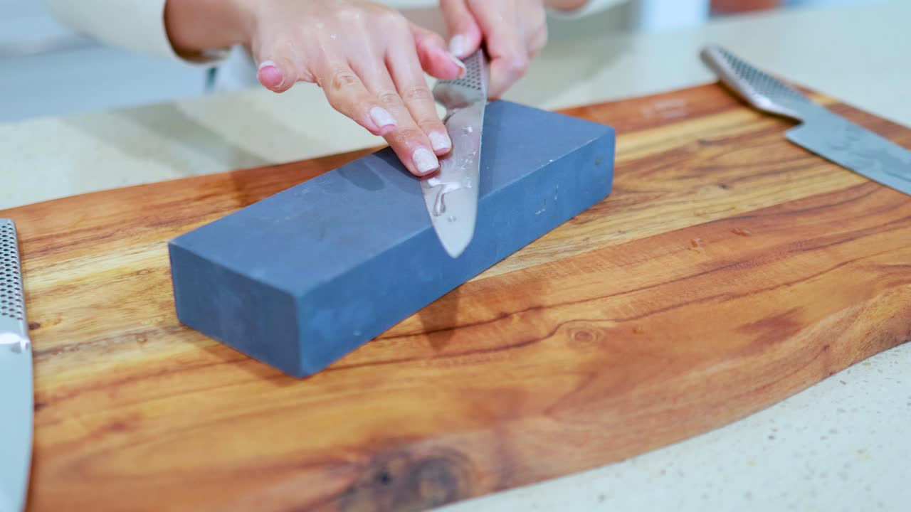 Hands methodically sharpen a chef’s knife on a rectangular whetstone atop a wooden cutting board, under soft, even kitchen lighting with steady camera framing