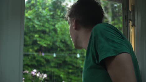 white man in green t-shirt looking out of door to rainy garden